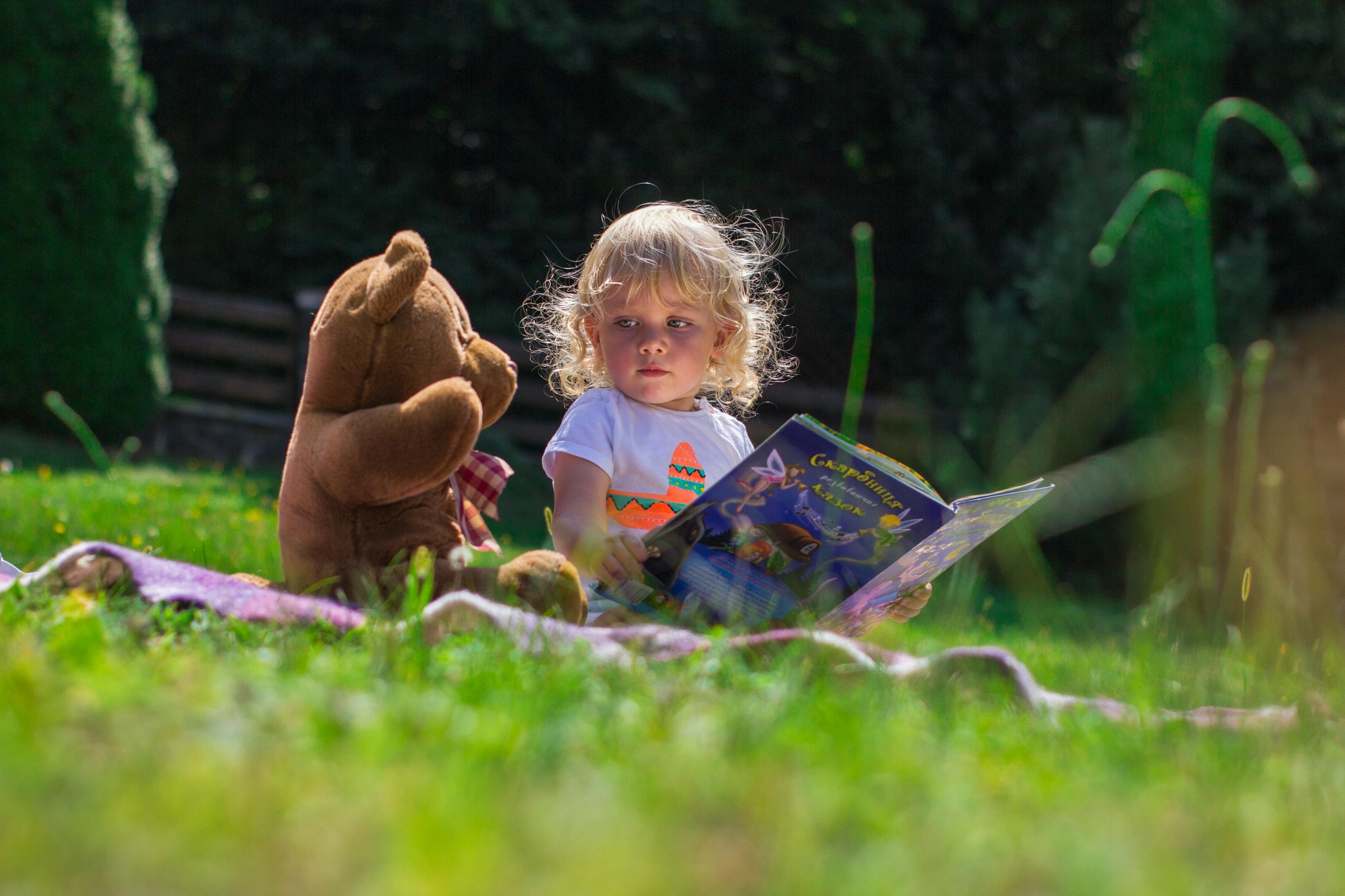 a little girl reading a story book to her teddy in the garden